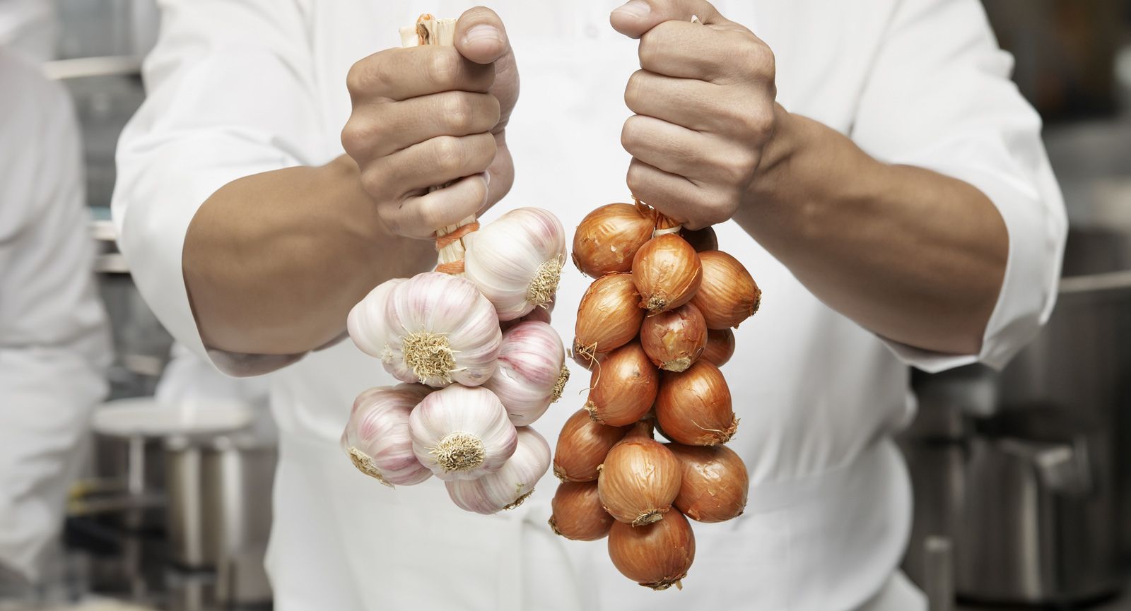 torso of person wearing chef coat holding a bunch of garlic and a bunch of onions