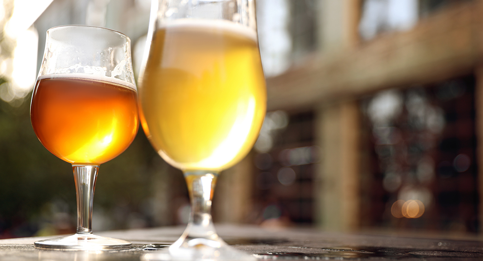 two stemmed glasses of beer with slight foam sitting on a table in sunlight