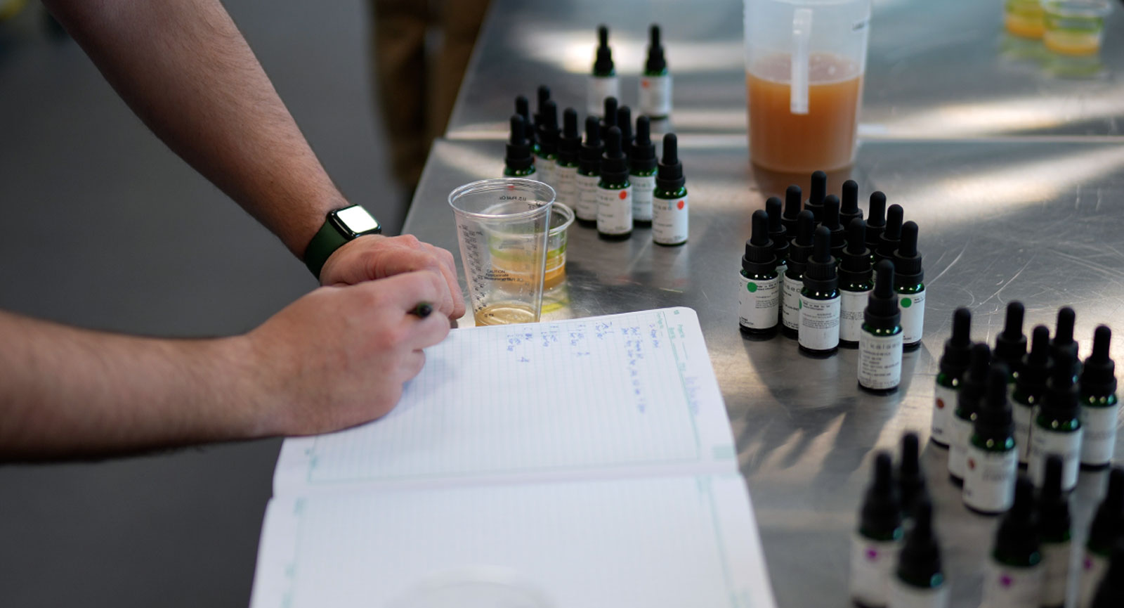 person in a brewery writing notes on a table covered with many glass dropper bottles of flavors