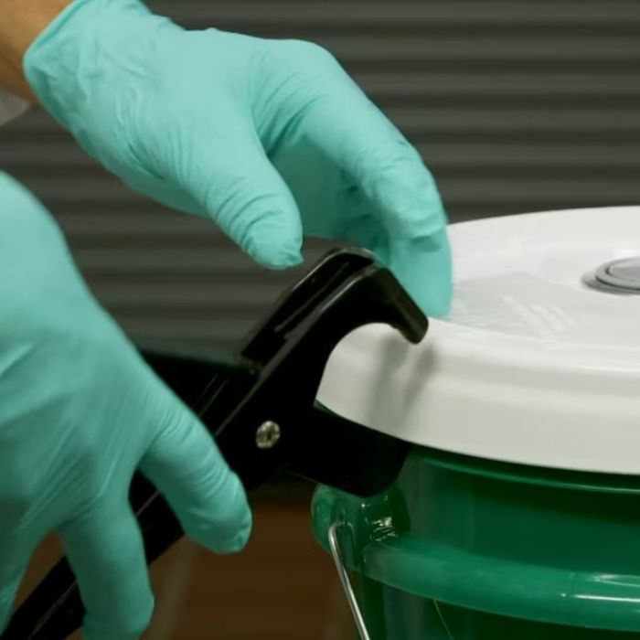 gloved hands using a pliers-like tool to lift a plastic lid from a container